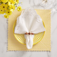 A white napkin with cutouts is held by the Kim Seybert Bois Napkin Ring in Brown/Gold and placed on a yellow-striped plate atop a woven yellow placemat, next to flowers and glassware.