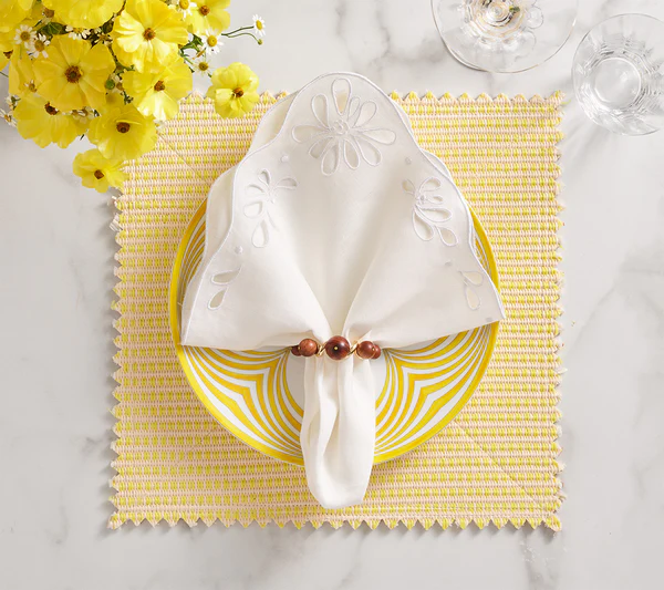 A white napkin with cutouts is held by the Kim Seybert Bois Napkin Ring in Brown/Gold and placed on a yellow-striped plate atop a woven yellow placemat, next to flowers and glassware.