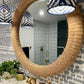 The Yuri Rattan Mirror by Made Goods hangs above a sink with gold fixtures; patterned wallpaper, pendant lights, and a potted plant complete the bathroom decor.