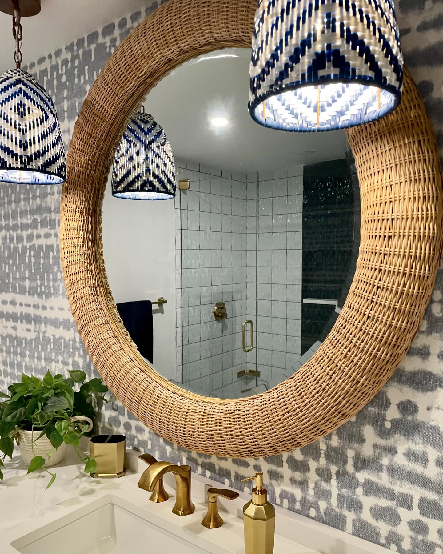The Yuri Rattan Mirror by Made Goods hangs above a sink with gold fixtures; patterned wallpaper, pendant lights, and a potted plant complete the bathroom decor.