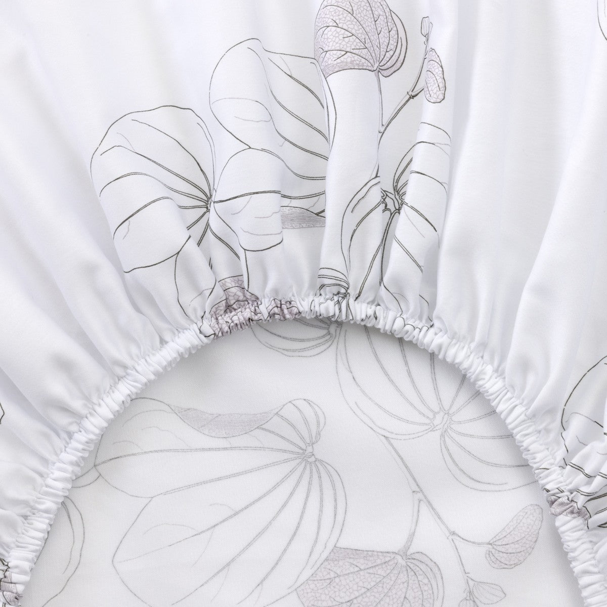 Close-up of a white fitted sheet from the Yves Delorme Orchidee Bedding Collection, made of organic cotton sateen with a subtle gray leaf pattern and elastic edging—an elegant touch to any floral bedding or French bed set.