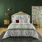 Yves Delorme Botanic Bedding Collection featuring a colorful floral and bird pattern on white linens, accented by matching pillowcases and a dark red cushion.