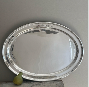 A Vintage Grand Platter from the Dorchester Hotel by Hôtel Silver leans against a light gray wall on a surface, with a single green pear beside it.