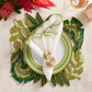 A modern table setting features a white plate on a green patterned placemat, a scalloped napkin with the Kim Seybert Palmera Napkin Ring in Gold, two glasses, and vibrant accents of tropical leaves and flowers.