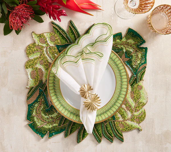 A modern table setting features a white plate on a green patterned placemat, a scalloped napkin with the Kim Seybert Palmera Napkin Ring in Gold, two glasses, and vibrant accents of tropical leaves and flowers.