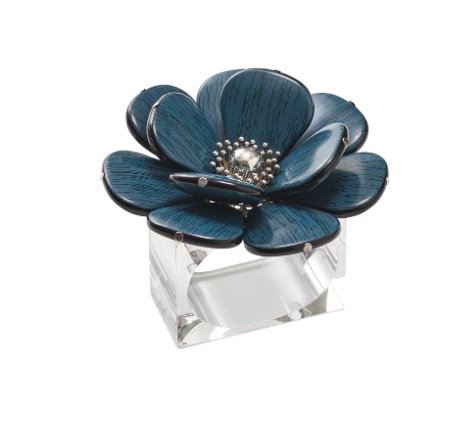 The Begonia Napkin Ring by Kim Seybert features a decorative blue and black floral design with silver accents, reminiscent of an intricate sculpture, mounted on a clear rectangular base.