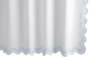 A Matouk Mirasol Shower Curtain, Blue, with a white diamond pique design, wavy scalloped edge, and light blue trim hangs vertically, combining elegance with the assurance of OEKO-TEX® certification.