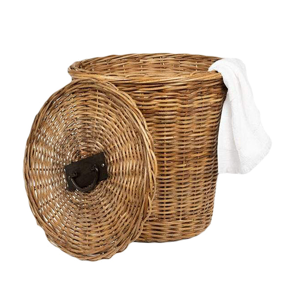 The Pigeon & Poodle Lamia Hamper, crafted from natural rattan, is a round wicker laundry basket with an open lid, adorned with iron handles and featuring a white cloth draped over the edge.