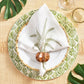 A table setting featuring a tropical leaf-patterned plate, bamboo-style charger, palm tree napkin, Kim Seybert Twist Brown/Gold Napkin Ring, wine glass, green leaf accents on a woven placemat, and gold inlaid brass details for elegance.
