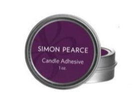 A small round metal tin labeled "Simon Pearce Candle Adhesive" sits slightly open in front of another tin—ideal for firmly securing candles to any candle base.