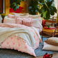 A bed with the John Robshaw Poseti Lotus Sheet Set in pink organic percale cotton, layered with a white quilt, assorted pillows, and a throw blanket. Plants and natural light fill the room; a pillow and red shoes rest on the floor.