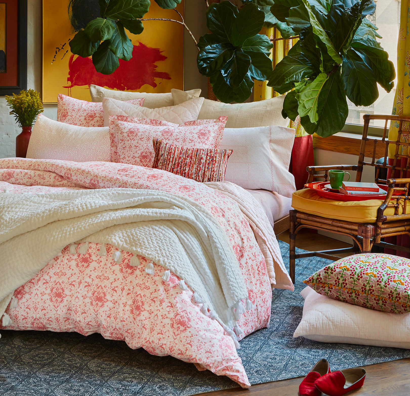 A bed with the John Robshaw Poseti Lotus Sheet Set in pink organic percale cotton, layered with a white quilt, assorted pillows, and a throw blanket. Plants and natural light fill the room; a pillow and red shoes rest on the floor.