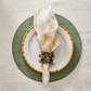 A white plate with a gold bamboo-patterned rim sits on a green charger, topped with a cream napkin held by the Kim Seybert Pavilion Brown/Gold Napkin Ring, featuring hand-carved mango wood and an inlaid brass edge.