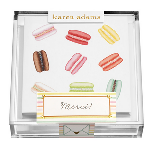 A clear box holds the Macarons Gift Enclosure Set by Karen Adams, featuring cards decorated with colorful treats and a "Merci!" card in front—perfect friendship cards to brighten anyone’s day.