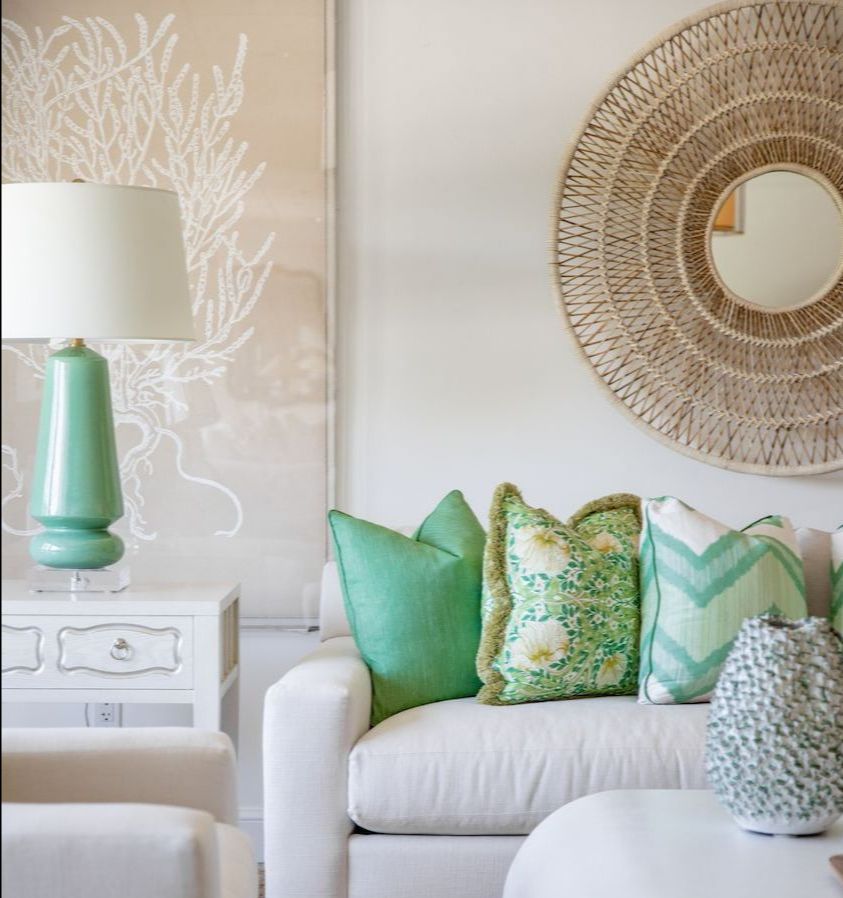 A stylish living room showcases a white sofa with handmade Chevron Green Ikat Pillows from Hive Custom Pillows, complemented by a green table lamp. A woven mirror and coral wall art complete the USA-inspired decor.