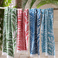 The Matouk Zebra Palm Beach Towel in Jungle, a patterned cotton terry towel, hangs on a wooden rod surrounded by tropical leaves and light wooden walls.