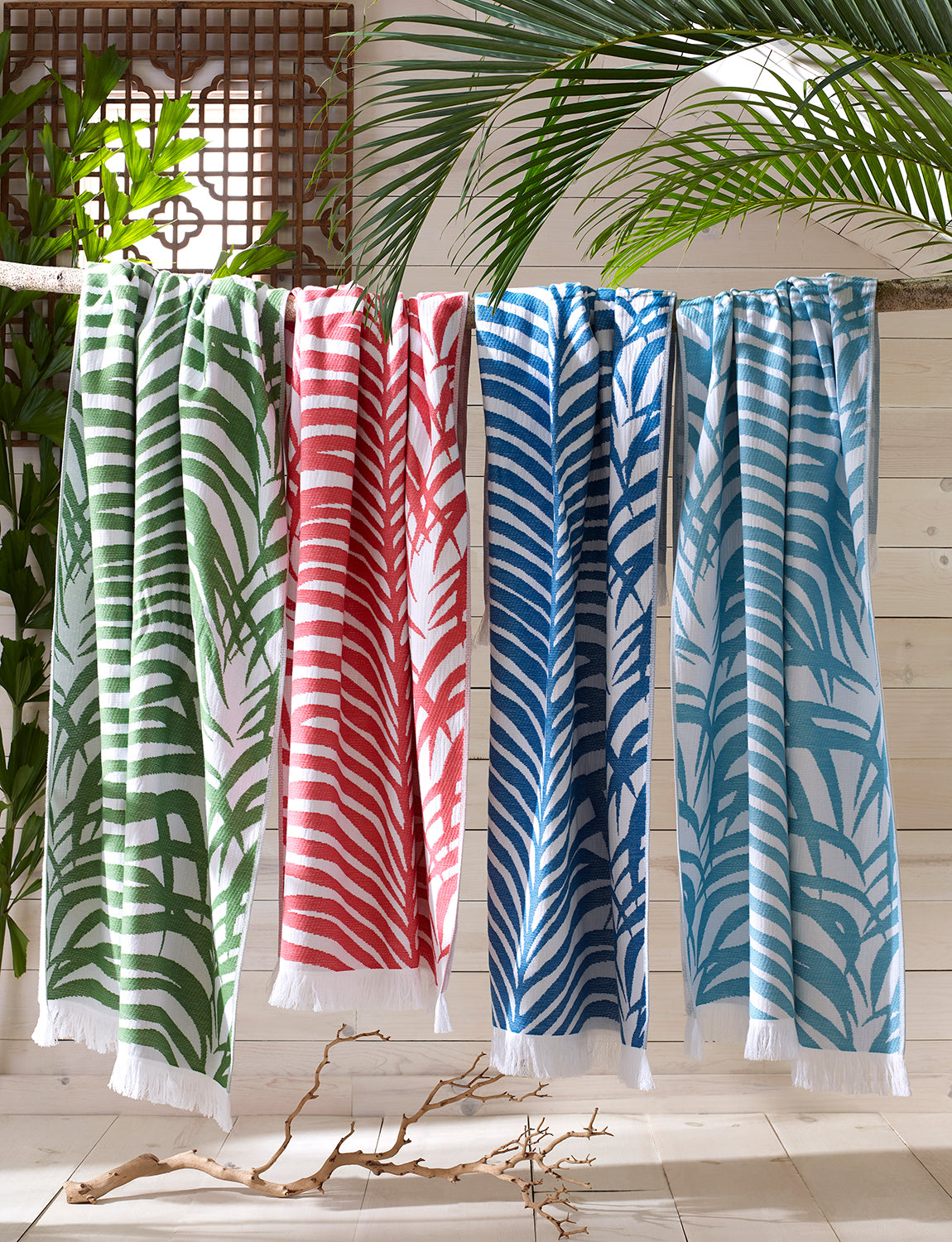 The Matouk Zebra Palm Beach Towel in Jungle, a patterned cotton terry towel, hangs on a wooden rod surrounded by tropical leaves and light wooden walls.