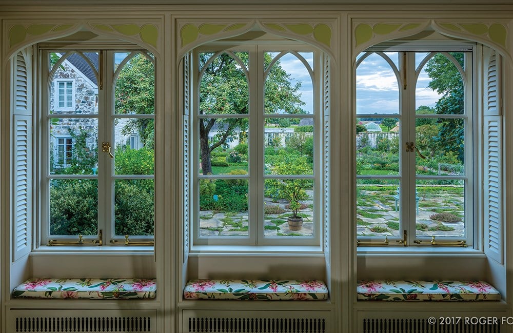 Through the three-paned window, a meticulously designed garden unfolds with trees and shrubs. Window seats feature floral-patterned cushions, echoing the elegance of Bunny Mellon from Common Ground's "Gardens of Bunny Mellon.