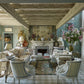 An ornate living room features patterned furniture, a decorative fireplace, large flowers in a vase, and framed art reflecting Pietro Cicognani's custom architecture. Light streaming through windows onto the checkered floor adds charm to the luxury home, echoing the style of Common Ground's "Pietro Cicognani: Architecture and Design.