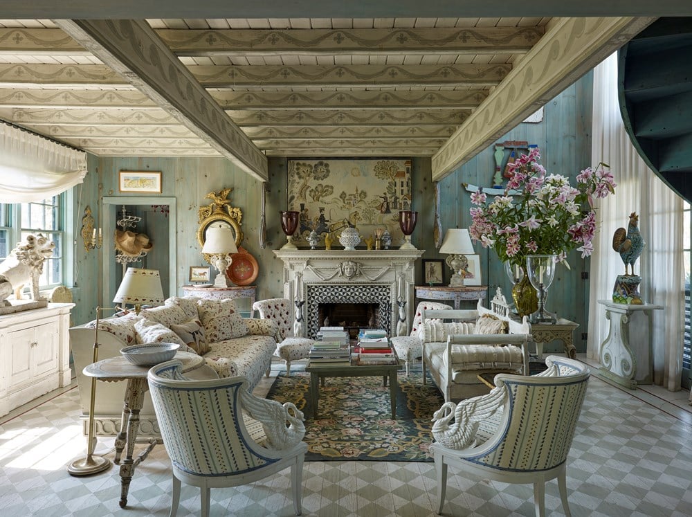 An ornate living room features patterned furniture, a decorative fireplace, large flowers in a vase, and framed art reflecting Pietro Cicognani's custom architecture. Light streaming through windows onto the checkered floor adds charm to the luxury home, echoing the style of Common Ground's "Pietro Cicognani: Architecture and Design.
