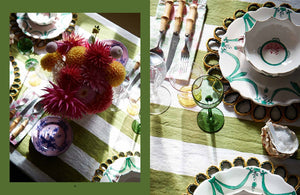 A table setting with colorful flowers in a vase, patterned plates, glasses, and cutlery arranged on a green and white striped tablecloth exudes a whimsical style reminiscent of A Kind of Magic: The Kaleidoscopic World of Luke Edward Hall by Common Ground.