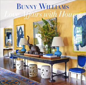 A stylish living room featuring yellow walls, blue accents, art, a console table with lamps and ceramics reflects Bunny Williams’ design style. Text: “Love Affairs with Houses” by Abrams.
