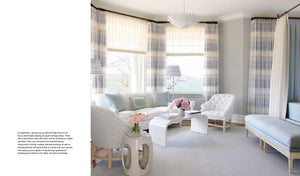 Discover elegance in "The Principles of Pretty Rooms" by Abrams, showcasing a refined interior with white and light blue decor, featuring chairs, a loveseat, side tables adorned with flowers, and large windows with striped curtains.