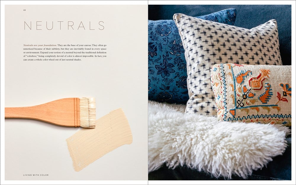 A beige-painted brush rests on a white surface near Penguin Random House’s "Living with Color," while patterned pillows and a white fur throw enhance the blue sofa, showcasing stylish home decor.