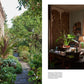 A tranquil pathway from the Common Ground's Kinfolk Garden meanders past the rustic exterior of the house. Within, a snug room features wooden furniture, lively plants, a lamp, and a tapestry adorning the wall as sunlight pours through the window.