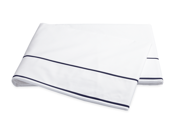White Matouk Ansonia Bedding Collection Egyptian cotton percale bedsheet with a navy blue border, folded neatly, isolated on a black background.