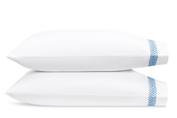 Two white pillows from Matouk’s Astor Braid Bedding Collection in Sky, each featuring a blue geometric embroidered edge, crafted from luxurious Ceylon percale.
