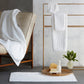 A cream chair, a tray with toiletries and flowers, and a gold rack displaying Matouk Auberge Monogram Bath Towels create an inviting guest bath inspired by Auberge style.