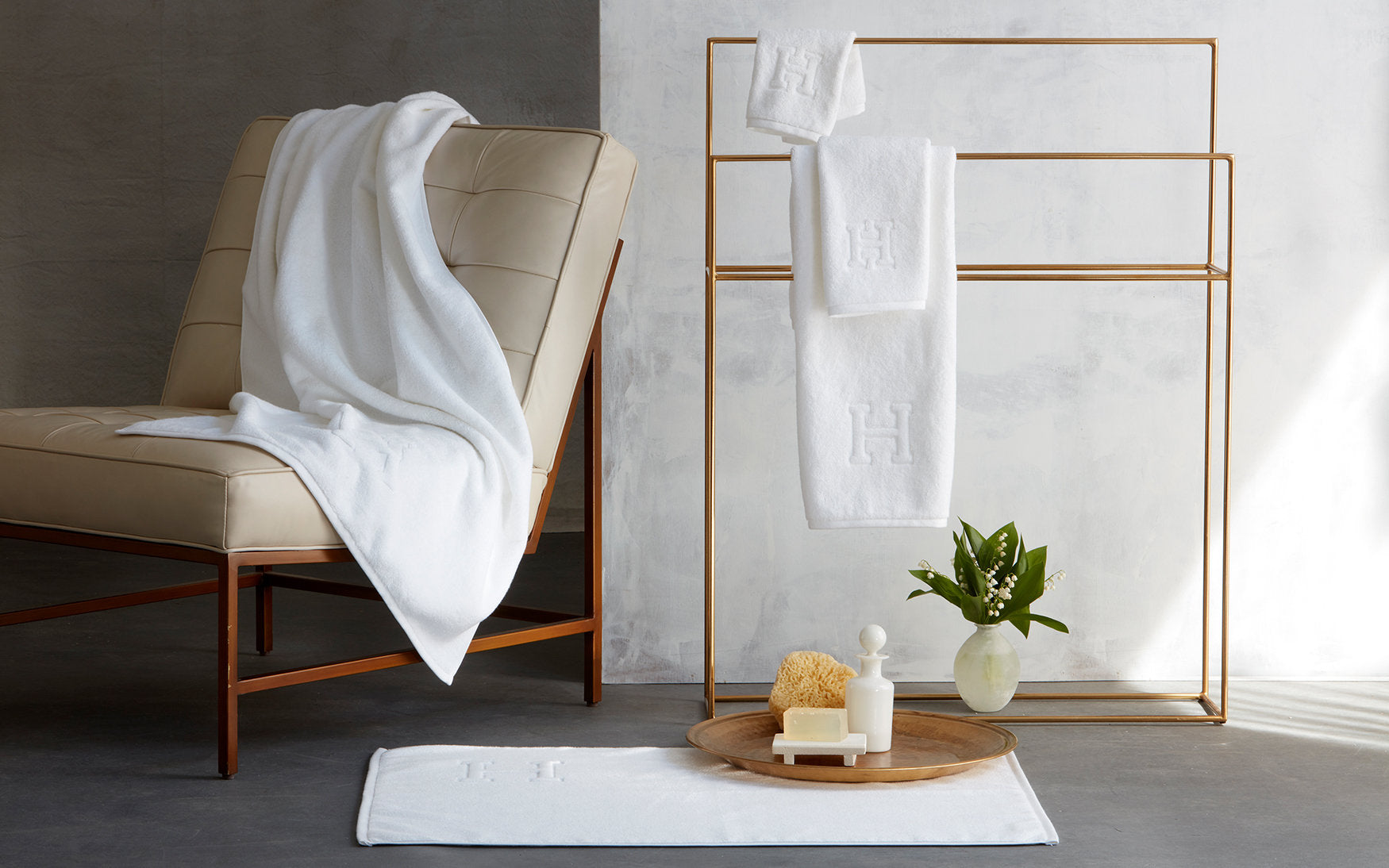 A cream chair, a tray with toiletries and flowers, and a gold rack displaying Matouk Auberge Monogram Bath Towels create an inviting guest bath inspired by Auberge style.