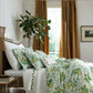 A neatly made bed with Matouk’s Pomegranate Bedding Collection in Citrus, several pillows, a potted plant by the bed, and golden-brown curtains covering tall windows in a bright bedroom.