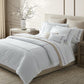 A neatly made bed with Matouk's Callista Bedding Collection in lilac and decorative pillows sits between two wooden nightstands in a bright, modern bedroom, each holding a lamp and a vase with branches.