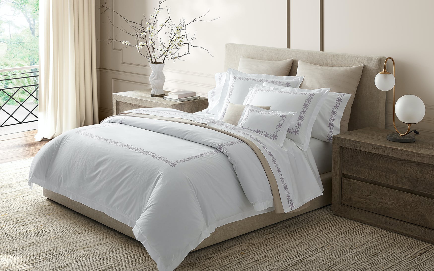 A neatly made bed with Matouk's Callista Bedding Collection in lilac and decorative pillows sits between two wooden nightstands in a bright, modern bedroom, each holding a lamp and a vase with branches.
