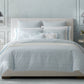 A modern bedroom with the Matouk Duma Diamond Bedding Collection in Sky, featuring light blue and white Egyptian cotton, two round side tables with lamps, a flower vase, and soft geometric accents.