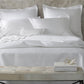 A neatly made bed features the Matouk Alba Bedding Collection in White, with pillows and a small bench at the foot holding books and glasses, all set against a concrete-style wall.