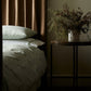 A bedside table with a vase of dried foliage stands next to a bed dressed in Matouk’s Duma Diamond Bedding Collection in Grass, made from OEKO-TEX® certified Egyptian cotton percale, with brown curtains in a dimly lit room.