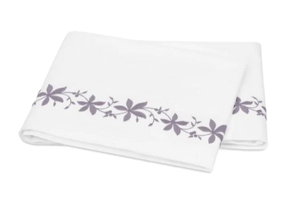 A folded white bed sheet from the Matouk Callista Bedding Collection, Lilac, features lilac floral embroidery along one edge and is crafted from luxurious Giza Egyptian cotton.