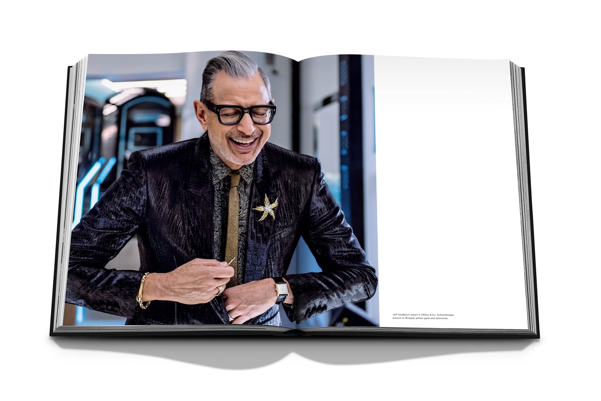 A man in glasses and a dark suit adjusts his sleeve while smiling in an open book spread, exuding luxury against a minimalistic background, featuring Diamonds: Diamond Stories by Assouline.