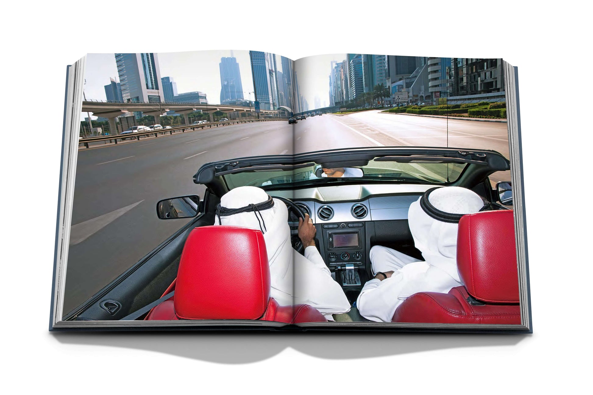 Two individuals in traditional attire are driving a car with red seats on an urban highway, detailed beautifully in the pages of Dubai Wonder by Assouline. Skyscrapers line the road in the background, showcasing the cultural diversity typical of Dubai tours.