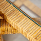 Close-up of a section of the Florida-Style Rattan Etagere by HIVE Home, Gift and Garden, featuring a wooden base with a glass surface. The wood, reminiscent of Palm Beach coastal style, boasts a ridged texture and natural finish. The glass edge is visible, showcasing a greenish tint.