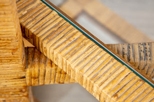 Close-up of a section of the Florida-Style Rattan Etagere by HIVE Home, Gift and Garden, featuring a wooden base with a glass surface. The wood, reminiscent of Palm Beach coastal style, boasts a ridged texture and natural finish. The glass edge is visible, showcasing a greenish tint.