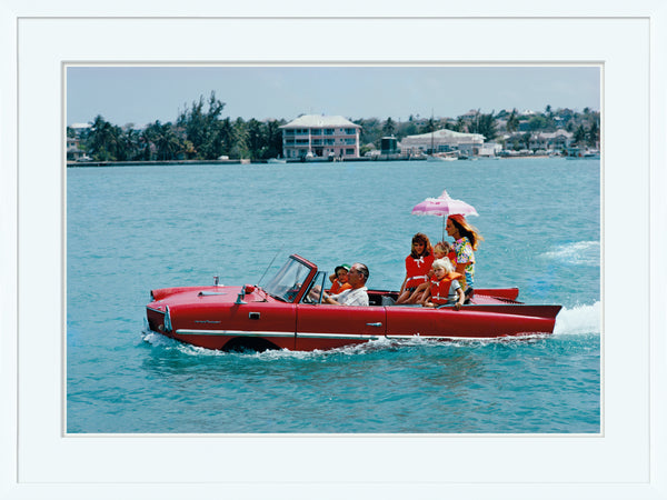 A family of four enjoying a sunny day on a red boat, with a child holding a pink umbrella, moving through calm blue waters near coastal buildings, reminiscent of Soicher Marin's "Sea Drive" by Slim Aarons By Getty Images Gallery, January 1, 1967.