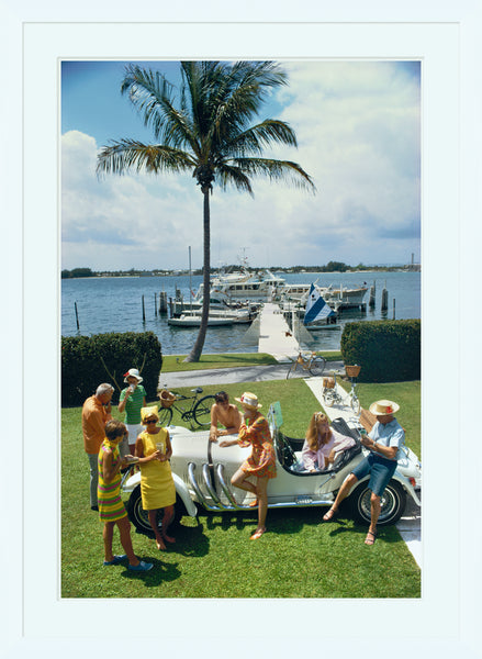 People in vibrant attire gather around a sleek white sports car on a grassy lawn near palm trees, with a dock and water in the background. This scene captures the timeless elegance of Slim Aarons' photography, as seen in Soicher Marin's "Palm Beach Society," April 1968 by Getty Images Gallery.