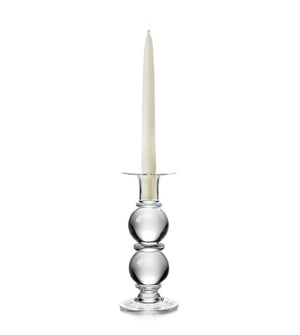 A white taper candle sits in the Simon Pearce Hartland Medium Candlestick, handmade by expert glassblowers with two round sections, shown against a plain white background.