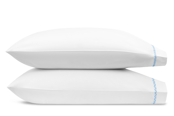 Two white pillows from the Matouk Hatch Bedding Collection in Sky, each with blue French knot edging, crafted from luxurious Ceylon percale, stacked horizontally against a plain background.