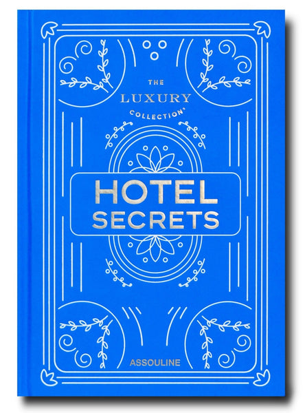 A blue hardback book from Assouline titled "The Luxury Collection: Hotel Secrets," featuring ornate white designs on the cover, reveals exclusive sights.