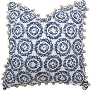 A handmade 22" x 22" pillow featuring a white background and a repeating blue circular pattern, adorned with beige pom-poms along the edges, the Tassel Inverted Medallion Navy Pillow by Associated Design.
